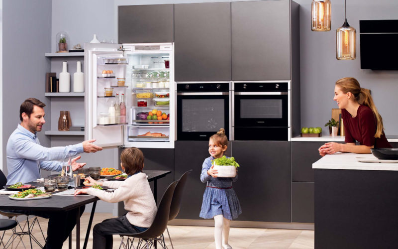 Family in their kitchen which showcases Samsung appliances. There are two ovens and a fridge