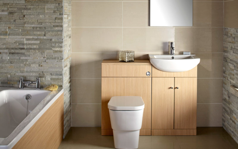Stone tiles by bathtub and window, with a white toilet and sink which is built into a wood cabinet